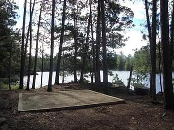 Square tent pad surrounded by trees and a scenic lake.